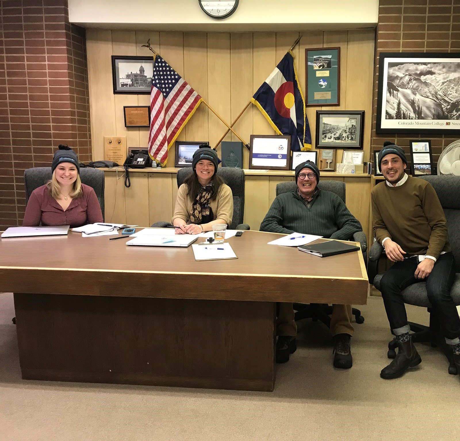 Four people wearing winter hats smiling sitting at a desk